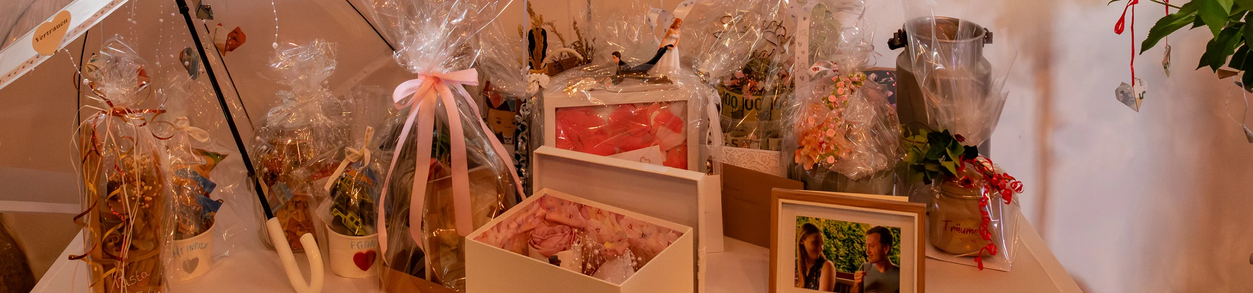 Table filled with wrapped gift baskets and ribbons, a framed photo, and decorative plants on a party table.